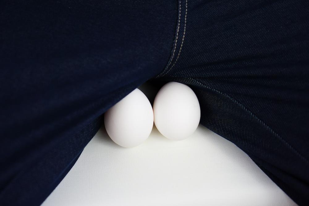 Close-up of two white eggs positioned on a white surface between the upper thighs of a person wearing blue jeans.